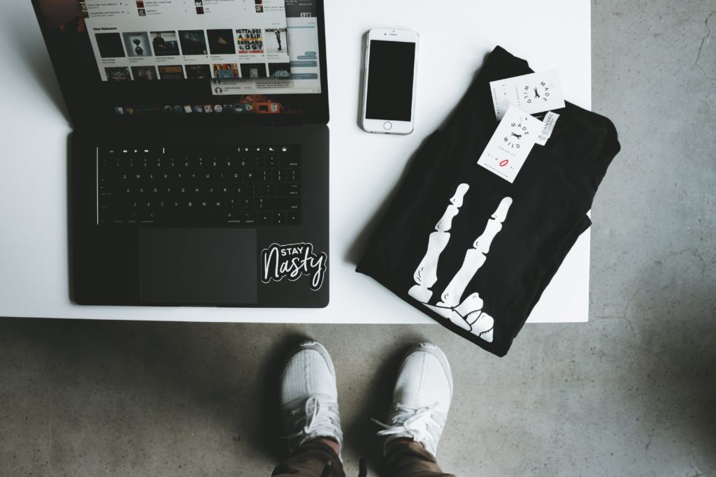Flat lay of a laptop with a stay nasty sticker, white iphone, black t-shirt with a skeleton giving the peace sign, and a person with white tennis shoes. 